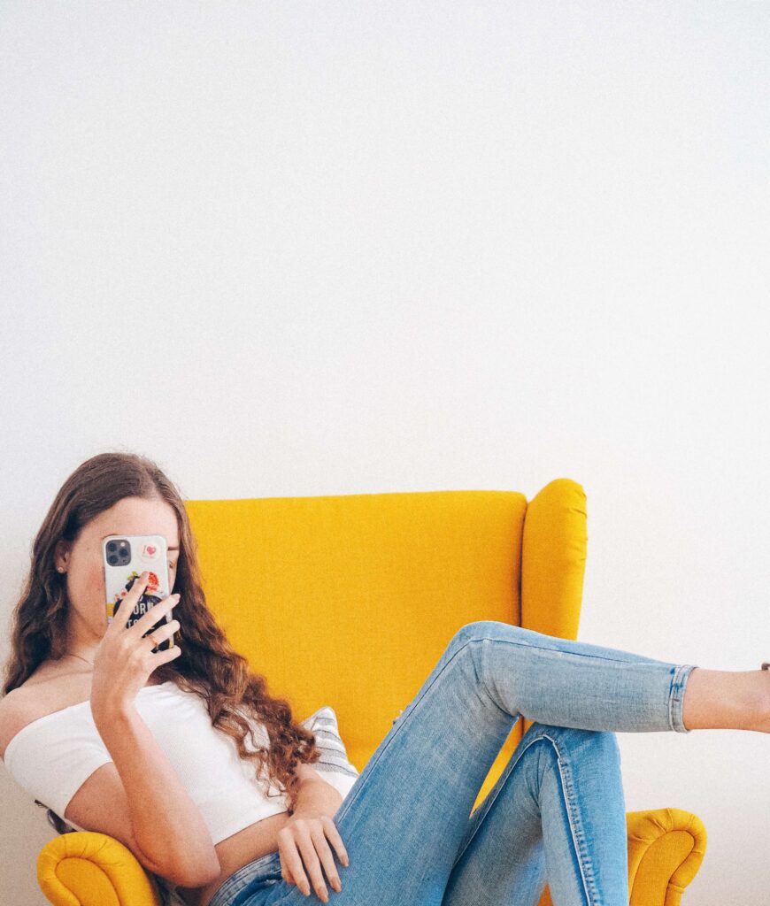 Foto einer jungen Frau die lässig auf einem gelben Lounge Sessel sitzt, die Beine übereinandergeschlagen, und in ihr Smartphone, das sie vor dem Gesicht hält, schaut – Levuro 